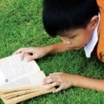 Boy reading a book on the grass to prepare for next school year.