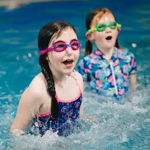 "Two young girls wearing swimsuits and goggles are in a swimming lesson.