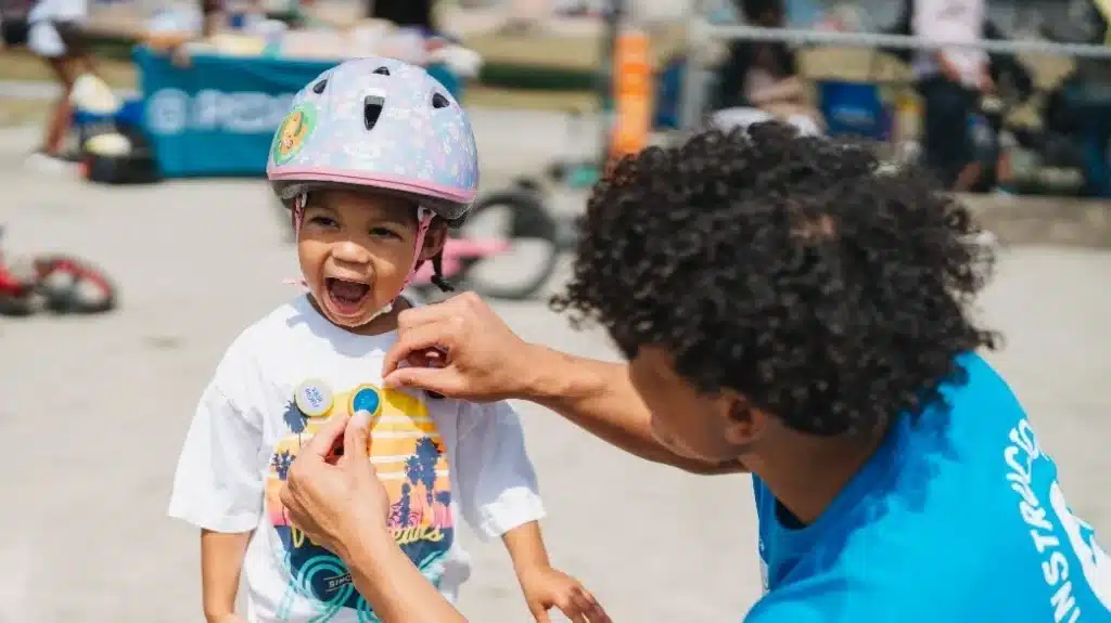 Male attaching a Pedalheads pin to the t shirt of a little girl with braids who is smilng. She is also wearing a helmet.